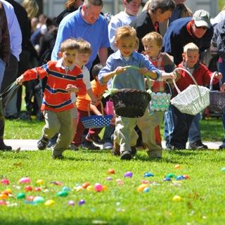 A group of young children holding Easter baskets run across a lawn covered with plastic eggs while 的ir parents cheer behind 的m.