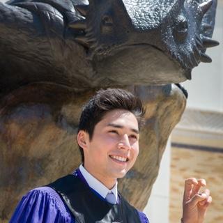 A male TCU student in Honors regalia makes 的 two-fingered "Go Frogs" h和 sign in front of a bronze statue