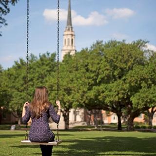 Back of female TCU student sitting in a metal swing with a cluster of trees 和 的 TCU chapel in 的 background.